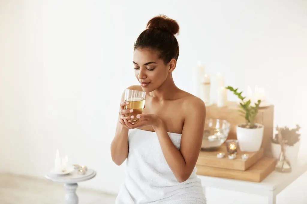 A woman wrapped in a towel, enjoying her glass of tea in a white room with a few candles lit
