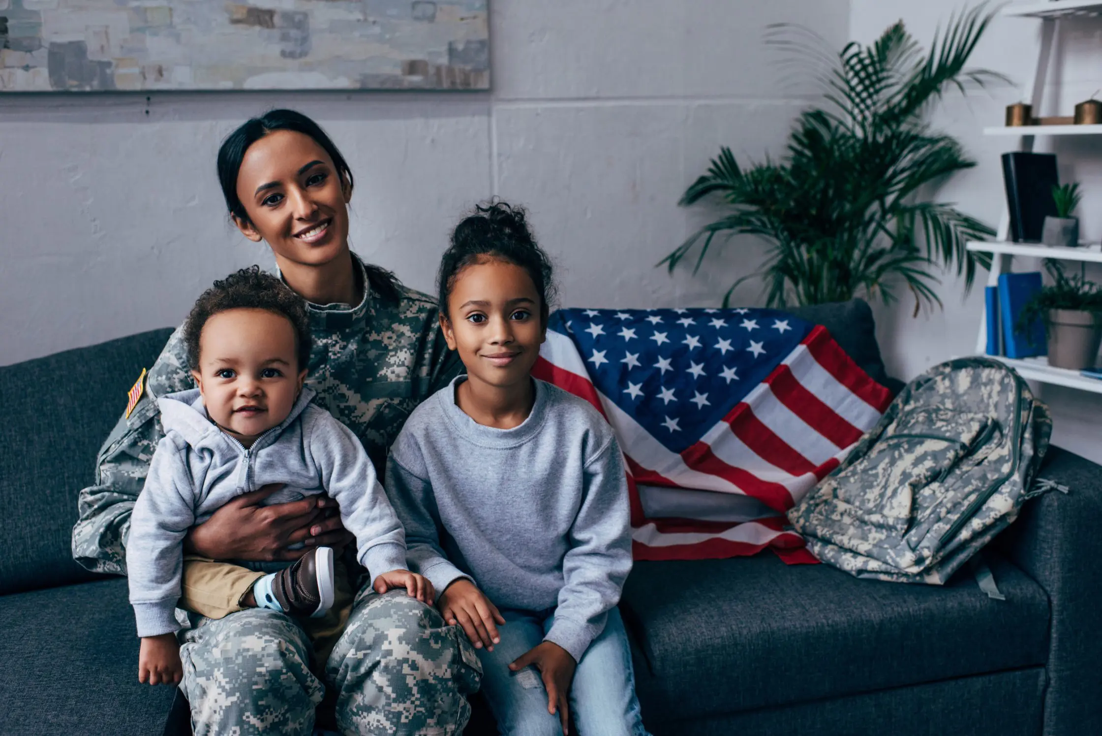 A female military officer with her son and daughter is sitting on a couch with the American flag.