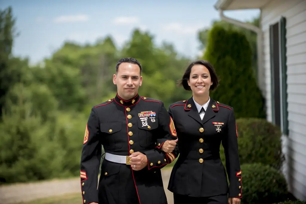 A marine couple standing side by side on their backyard surrounded by trees.