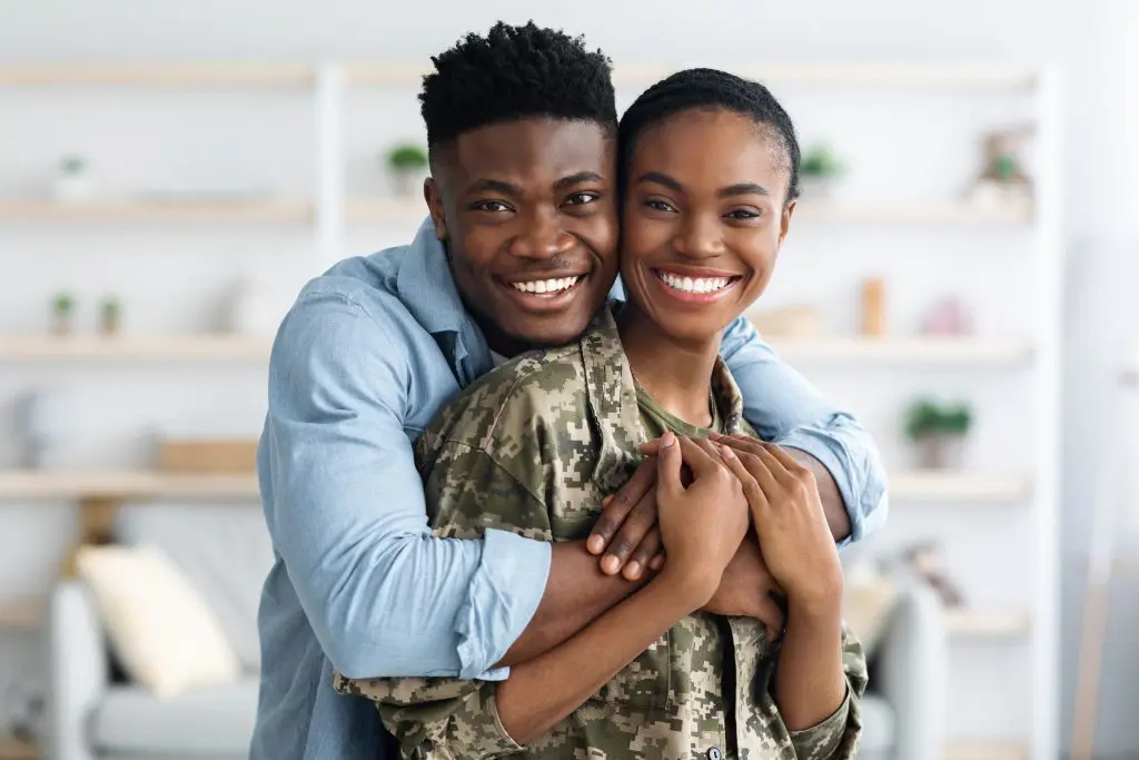 A military woman is embraced by her husband from behind, looking happy.
