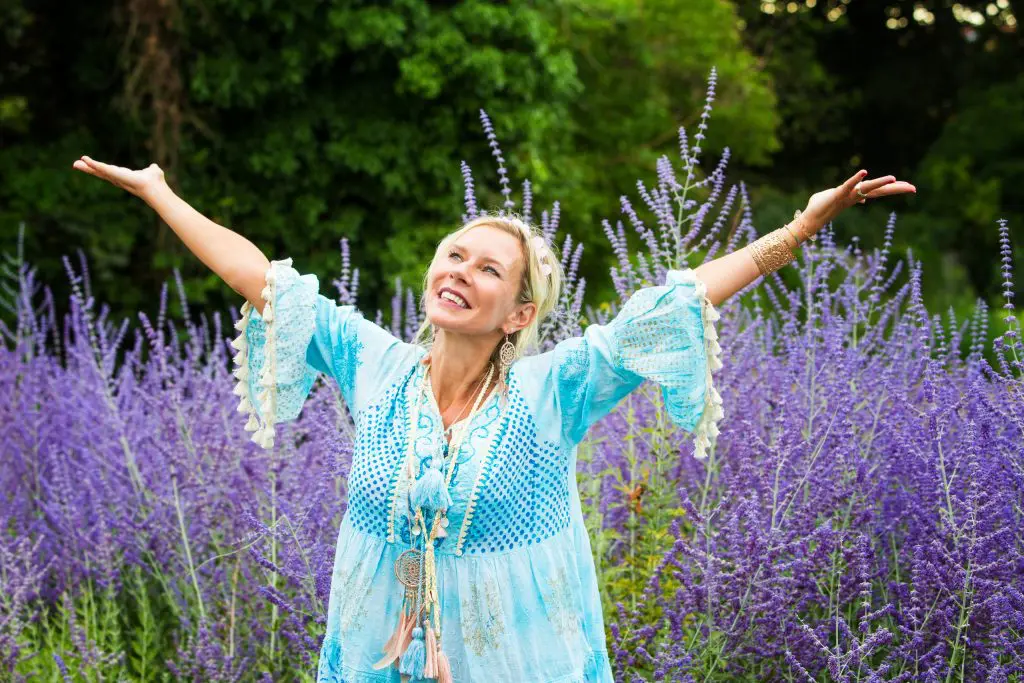 A woman in a blue dress is joyously raising both her arms up in the middle of a lavender field.