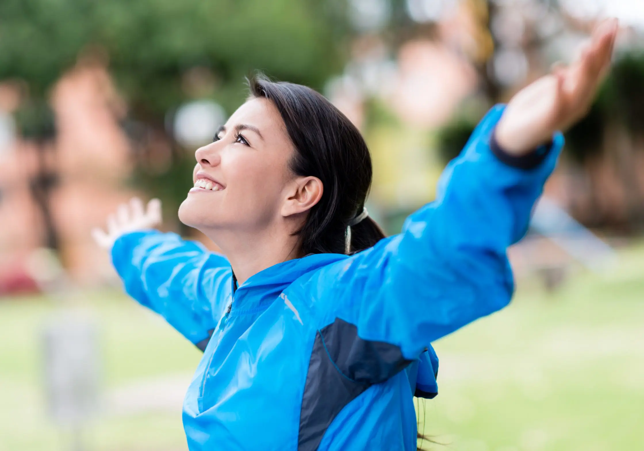 A woman in a blue jacket raises her hands above her head and smiles.