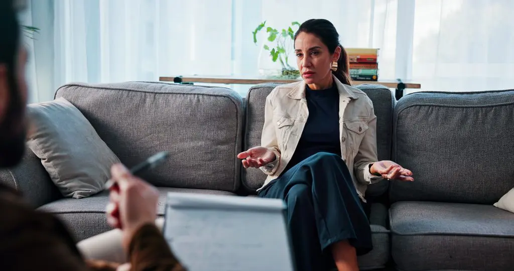 A woman sitting on a couch shrugs as the person sitting across from her points at her with a pen.
