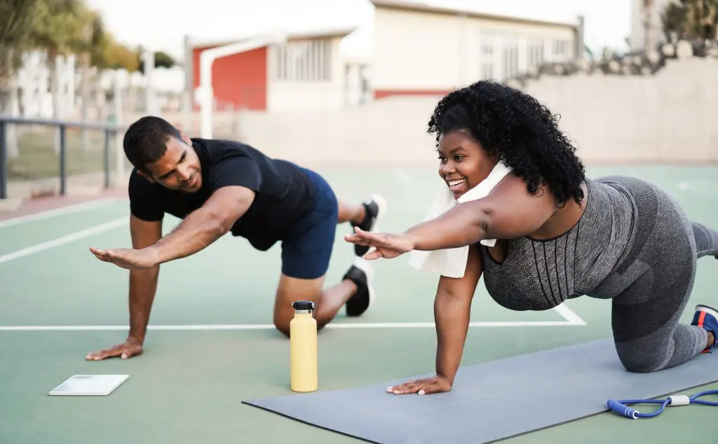 Two people wearing athletic clothing do yoga on a tennis court. One of them uses a yoga mat.
