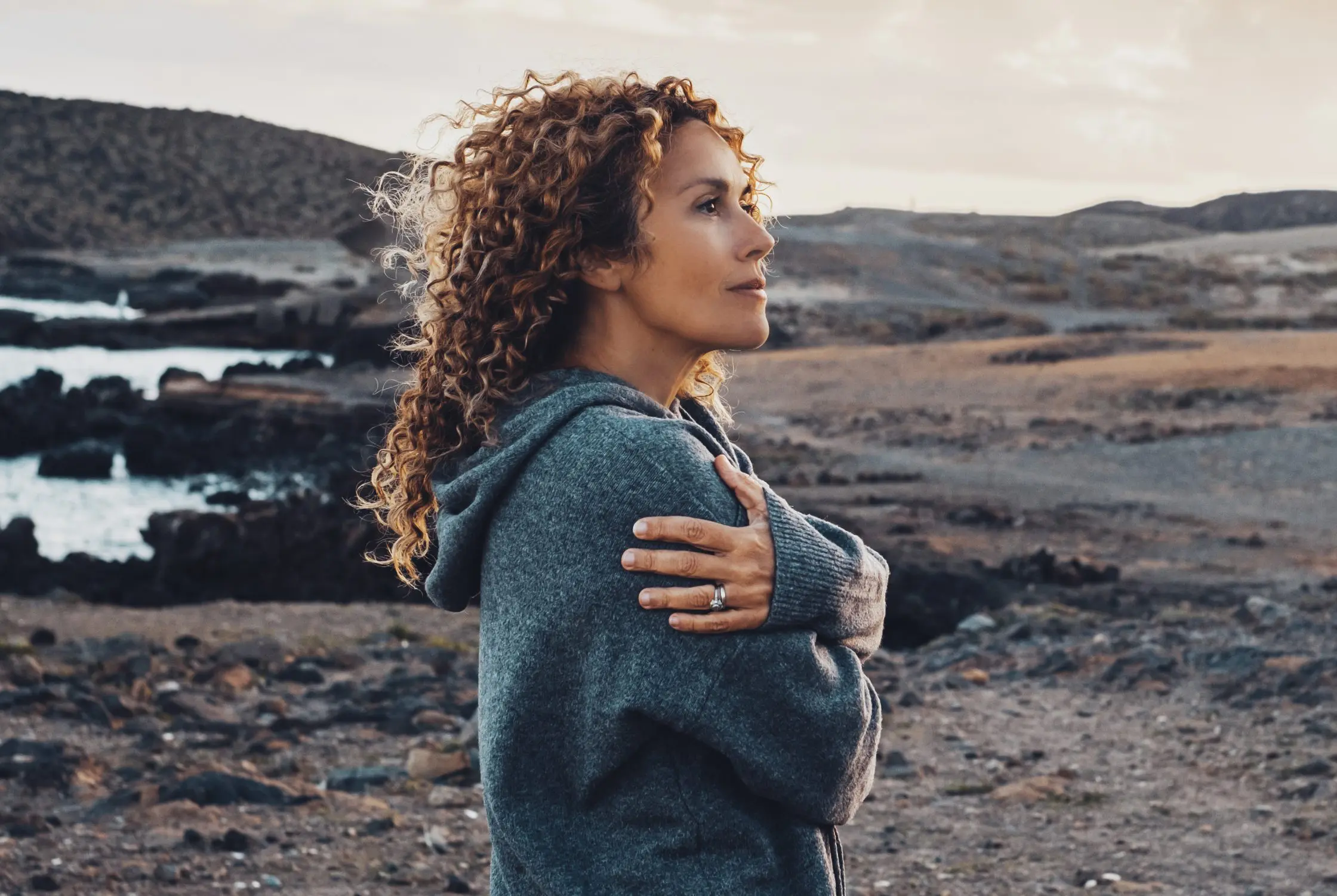 A woman standing outside near the ocean wraps her arms around herself in a hug.