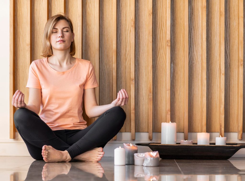 A woman wearing a pair of leggings and a peach shirt meditating with candles on her side