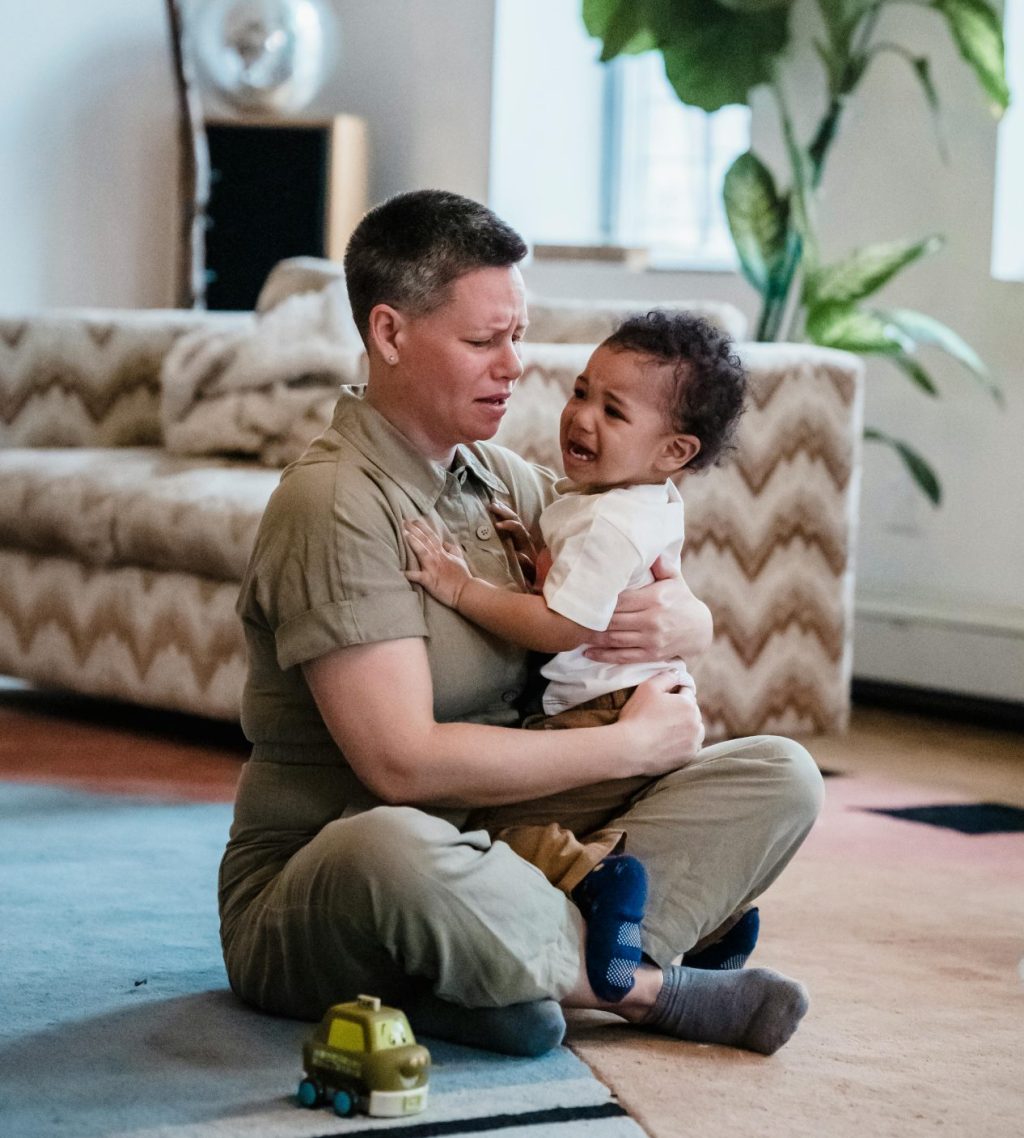 A woman looks worried because of the crying baby she is holding while sitting on the floor.