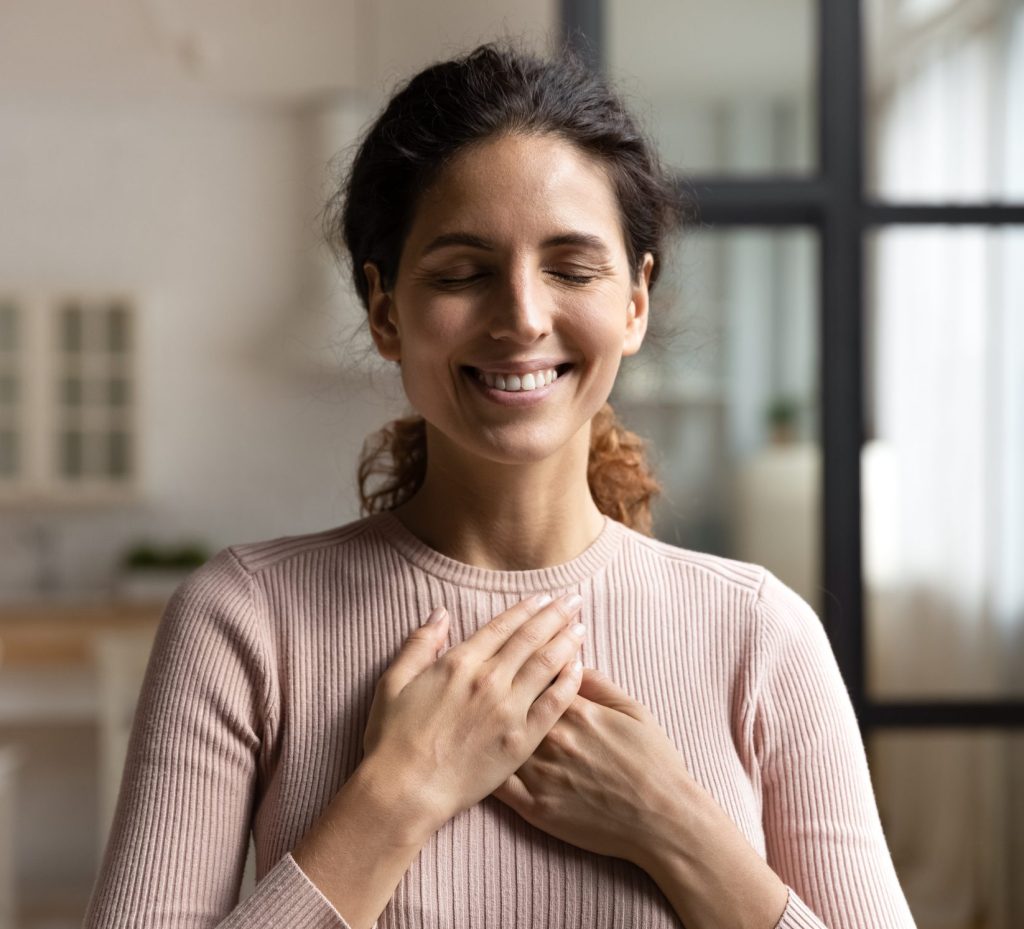 A woman wearing a ribbed shirt with both hands on her chest, her eyes closed, and smiling.